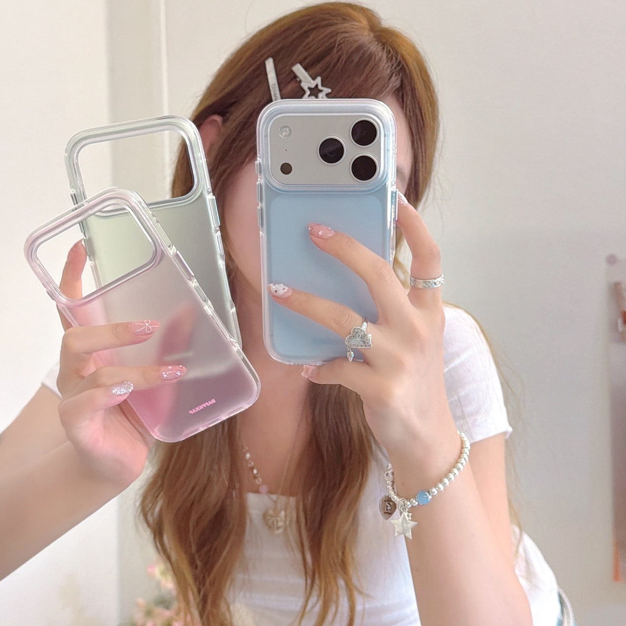 A young woman holding three aesthetic pastel phone cases in blue, pink, and green during a mirror selfie.