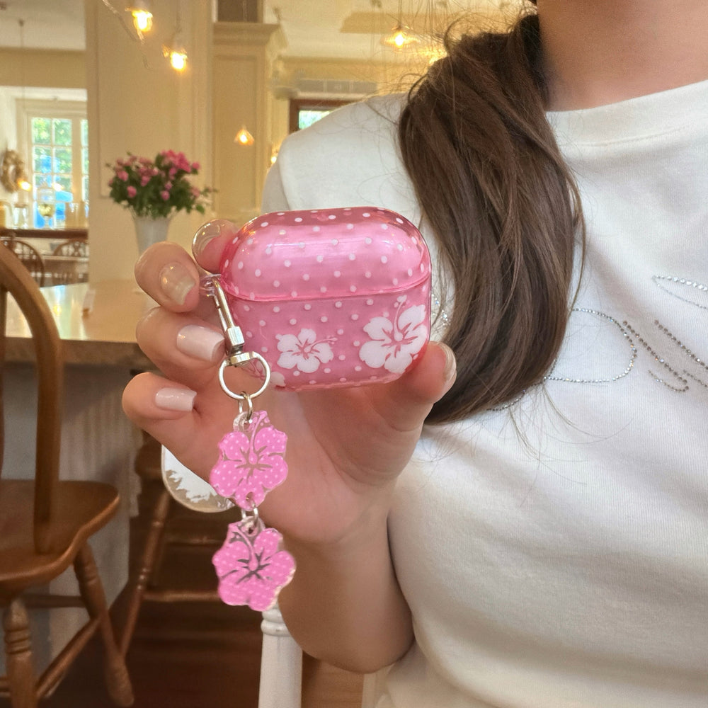 A girl holding a protective pink Hawaiian flower AirPods case with a dangling flower charm inside a cafe.