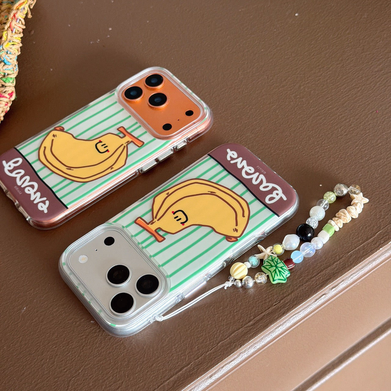 Two kawaii banana-themed iphone covers side by side on a brown table, one with a beaded wrist strap.