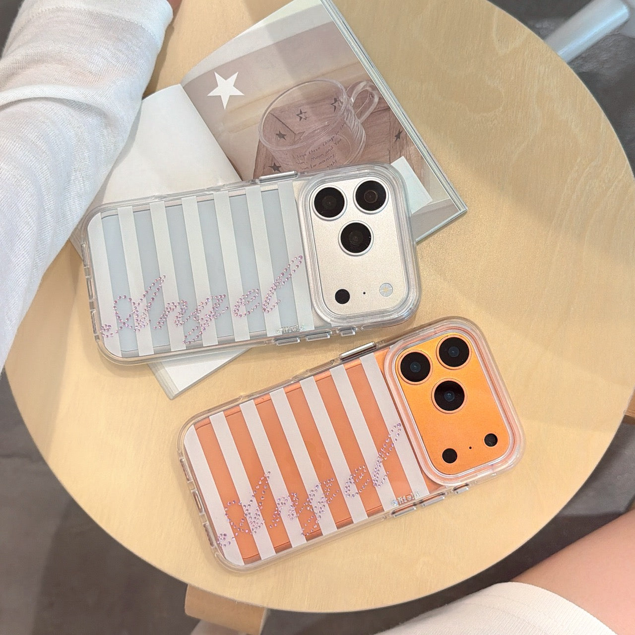 Two striped rhinestone iPhone cases in silver and orange colors displayed flat on a wooden table next to an open magazine.