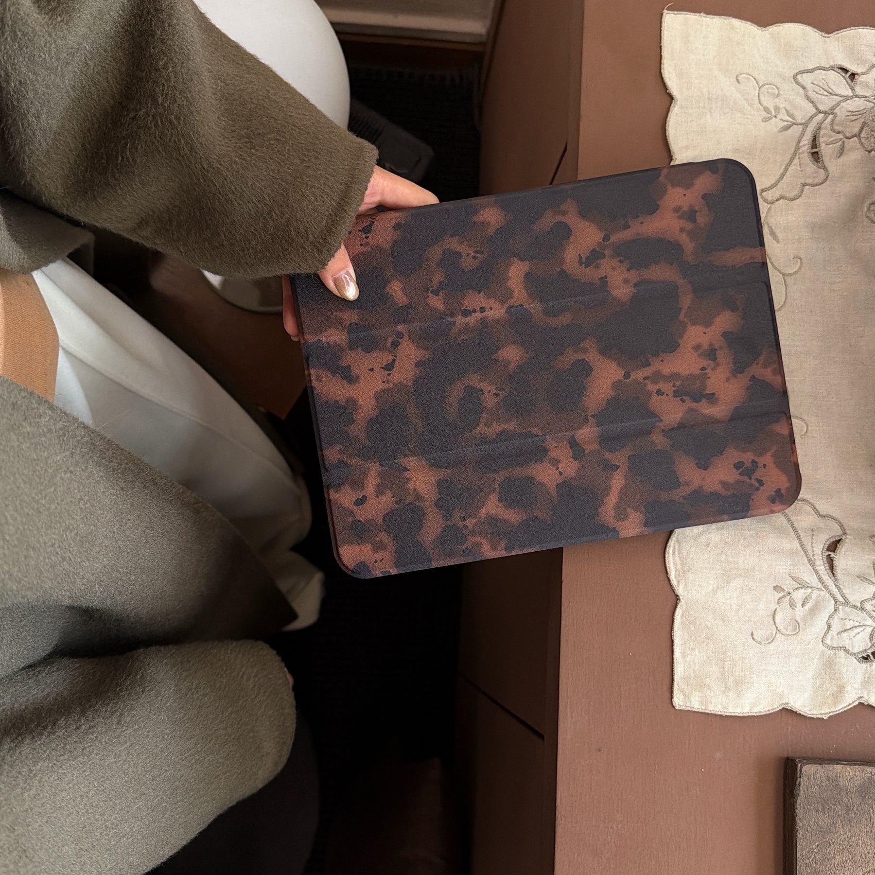 A person wearing an olive green wool coat holding a closed tortoiseshell tri fold tablet case over a brown desk with a vintage embroidered white lace mat.