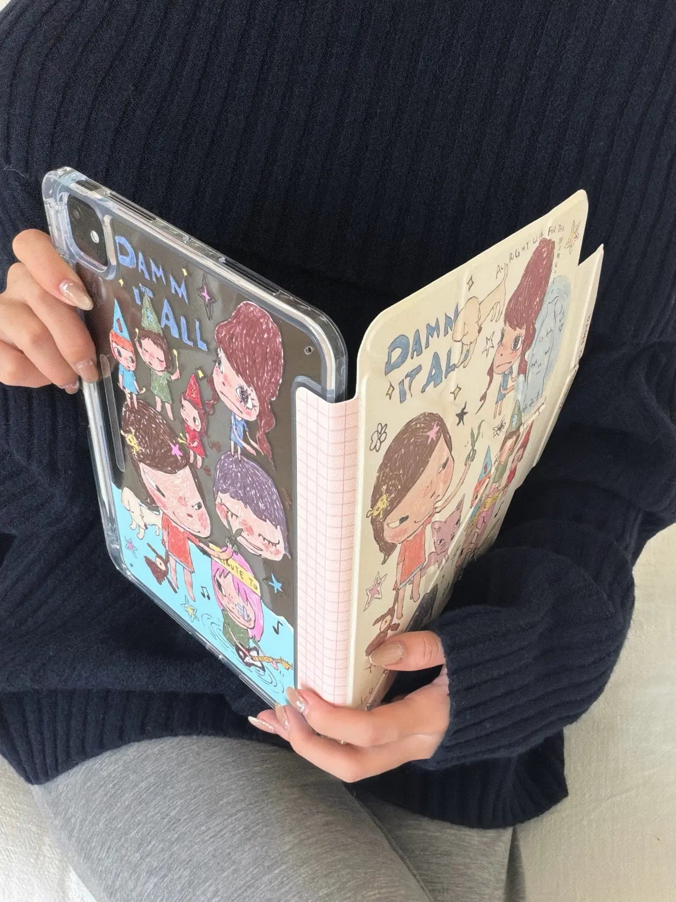 Side-by-side view of a Yoshitomo Nara iPad case being held open like a book, displaying the clear acrylic back with cartoon prints and a pink checkered spine.