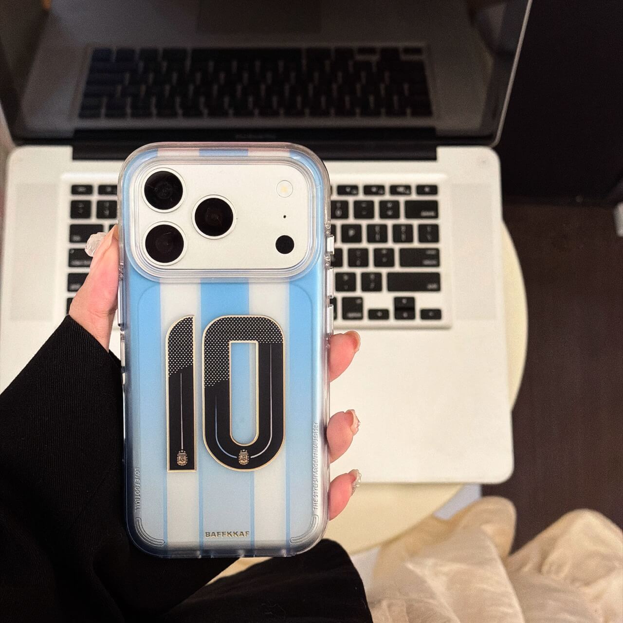 Hand holding a blue soccer jersey iPhone case with No.10 graphic near a laptop