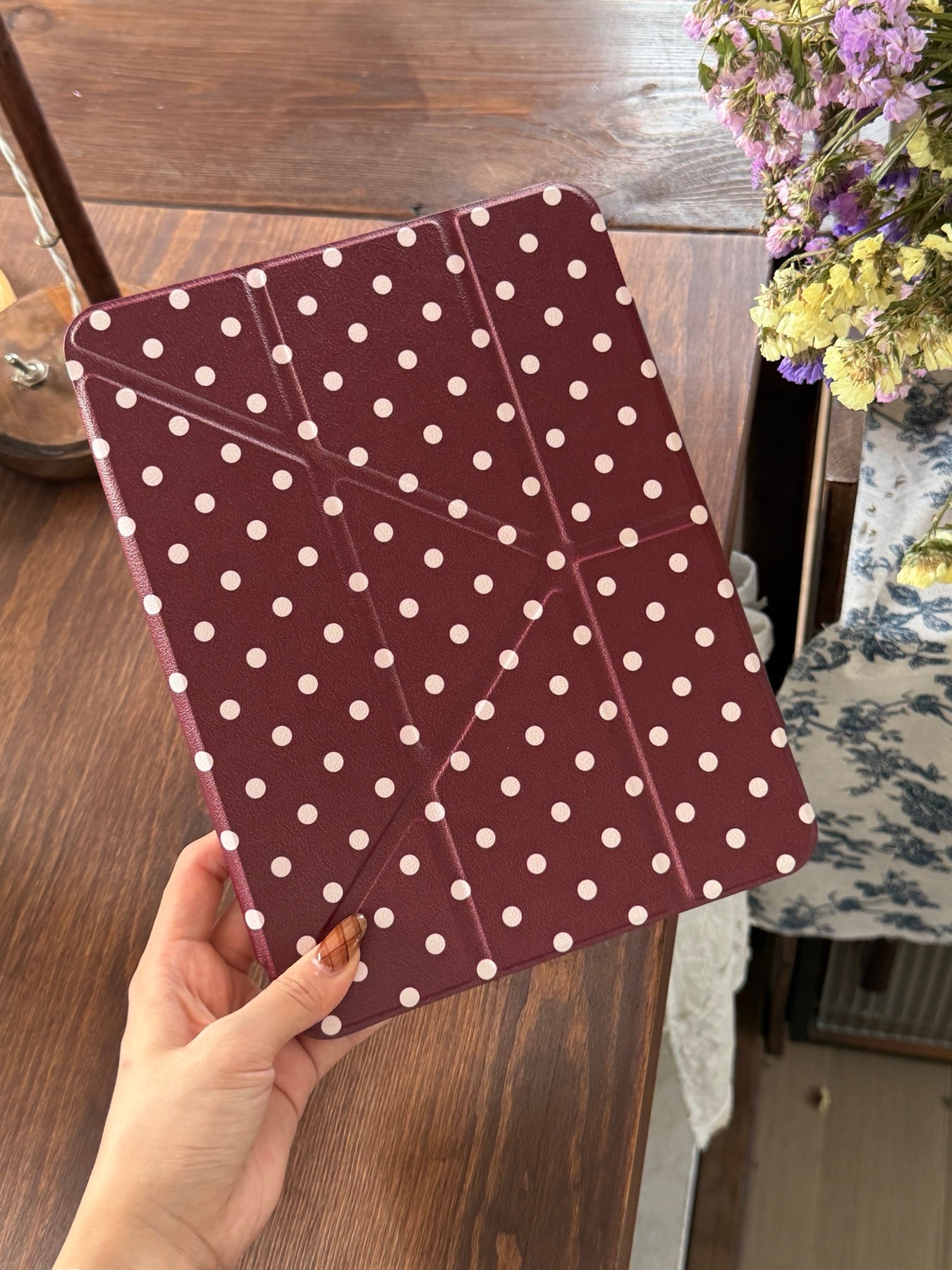 A hand holding a closed burgundy polka dot iPad case with origami geometric folding lines over a wooden table.