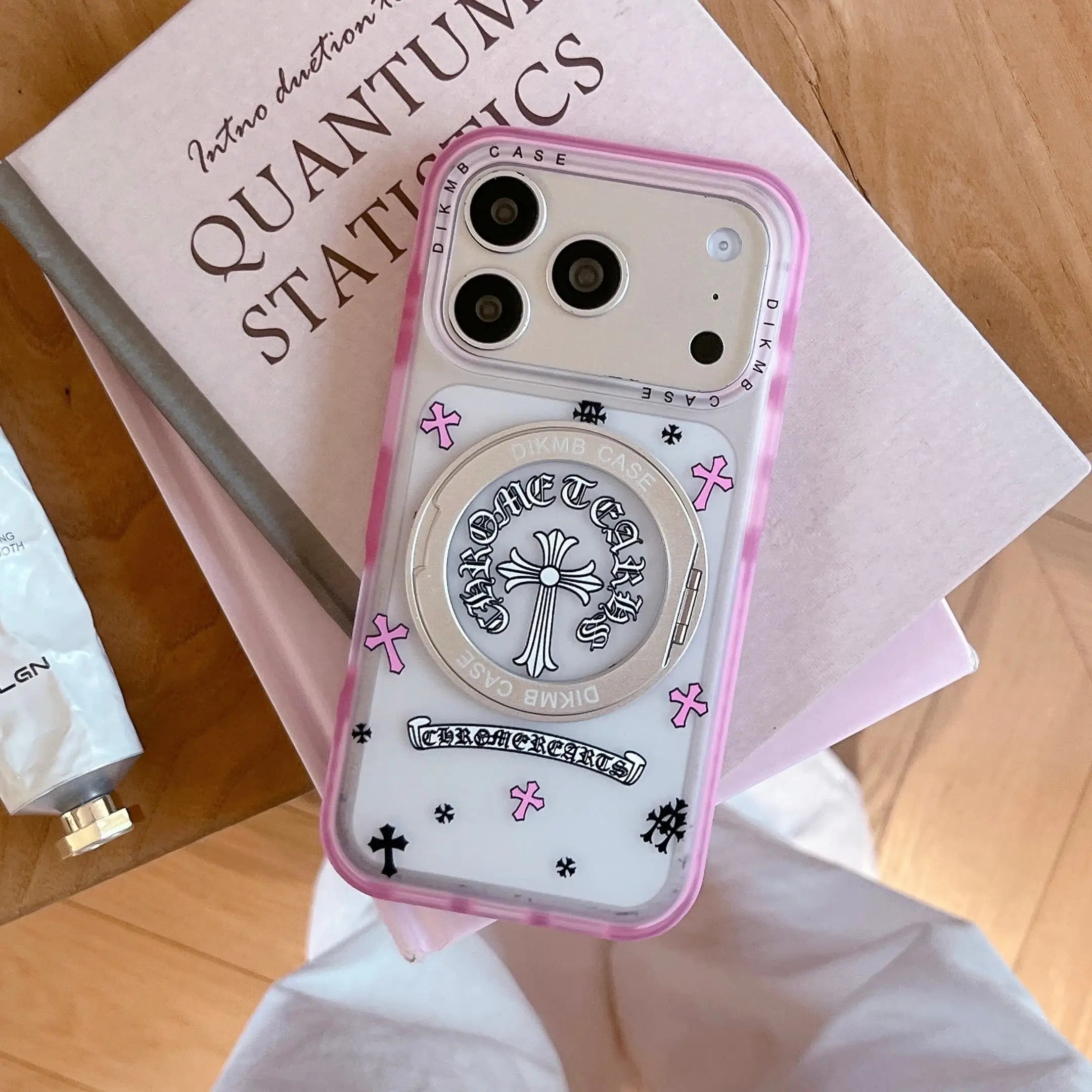 A Chrome Hearts inspired pink cross phone case with a built-in stand resting on a book.