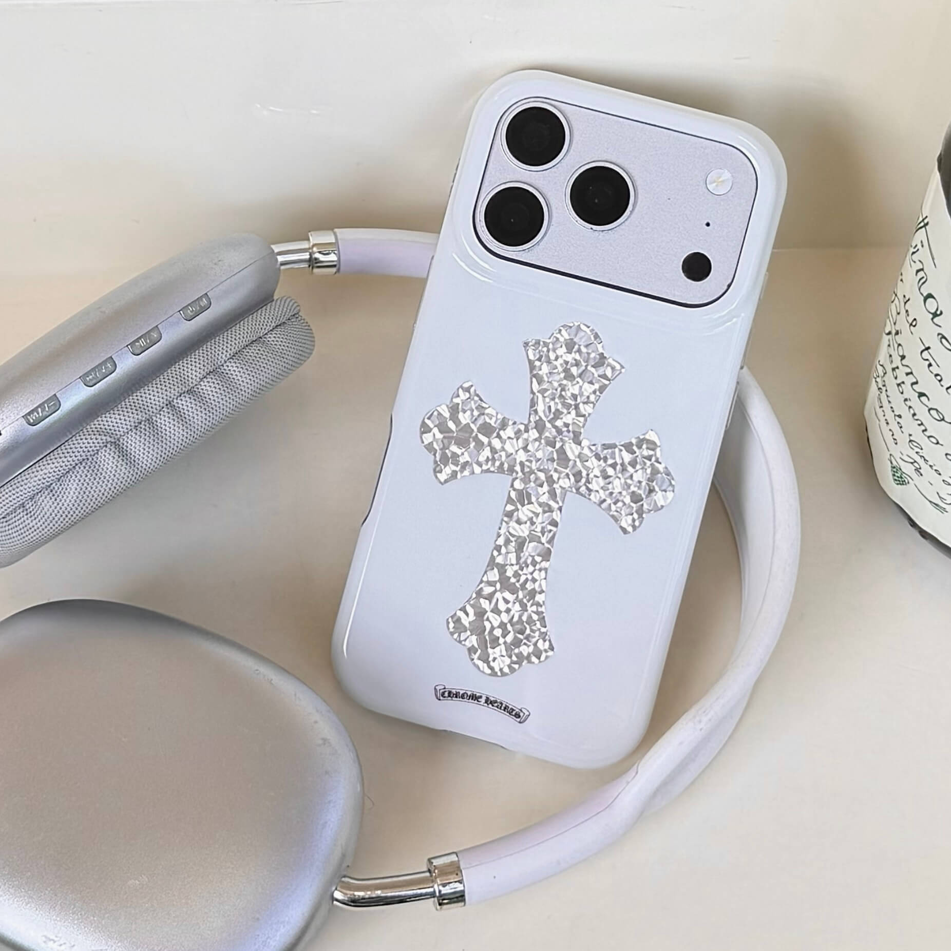 A white Chrome Hearts style silver cross iPhone case resting next to silver overhead headphones.