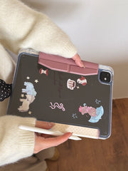 Rear view of an aesthetic clear back iPad case with printed cartoon graphics held horizontally, highlighting the pink magnetic closure clasp.