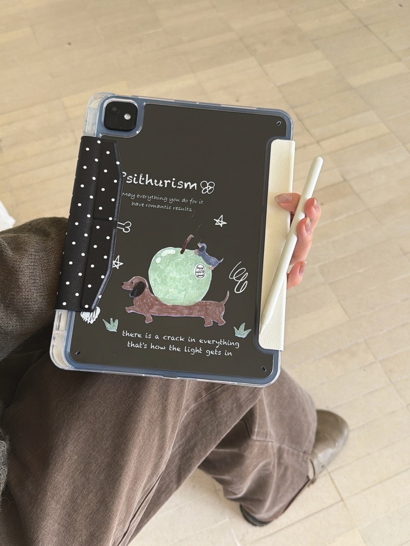 A person holding a clear back ipad case with pencil slot on their lap, displaying the funny sausage dog illustration on the back panel while holding a white Apple Pencil.