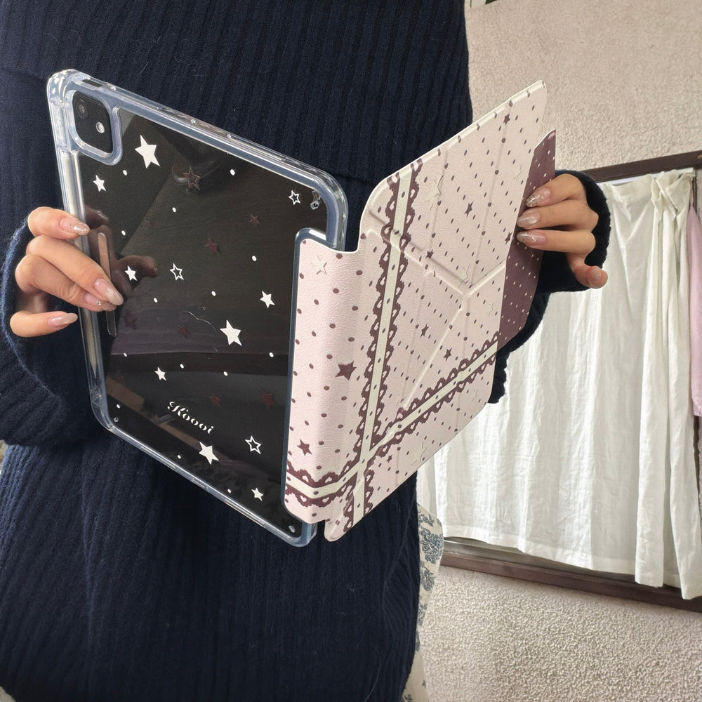 A person in a navy blue sweater holding an open coquette iPad case, showing the transparent starry acrylic back panel and heart lace front cover.