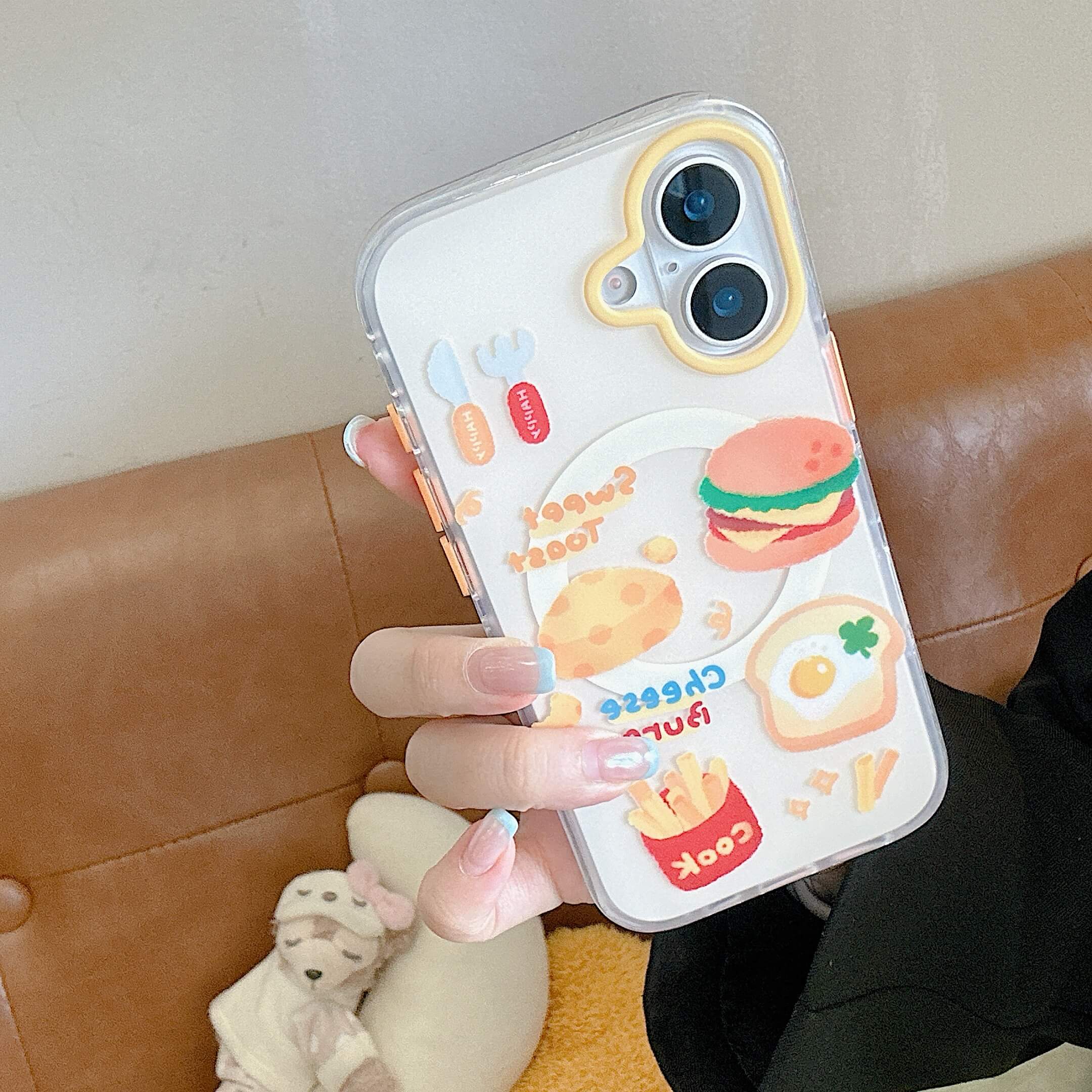 A hand holding the clear cartoon burger and fries phone case over a brown leather couch.