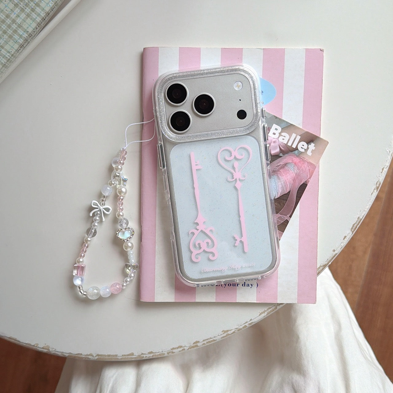 A clear glitter key phone case with a beaded strap laid out on top of a pink striped ballet notebook.