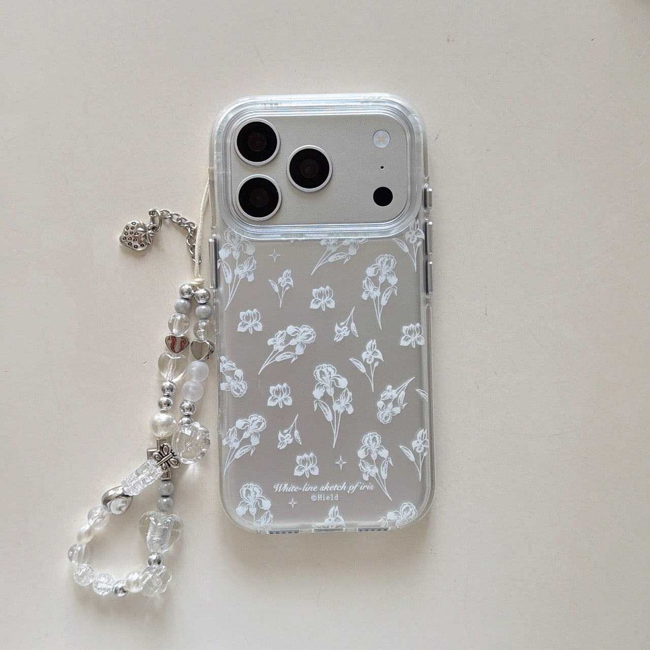 Product shot of a clear iris flower sketch phone case with an attached crystal and pearl charm strap on a white background.