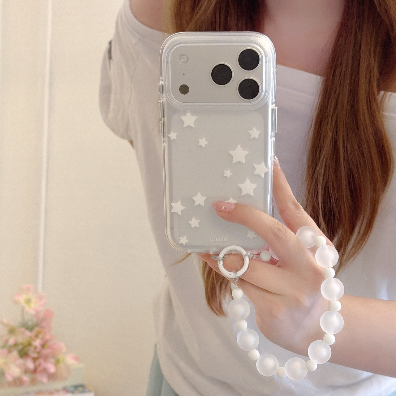 A mirror selfie of a girl holding a clear iPhone case with a white star pattern and a frosted beaded wrist strap.