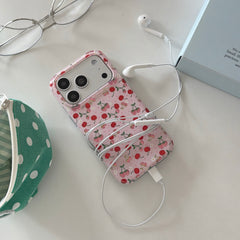 A coquette pink cherry phone case wrapped in white wired earphones next to a green polka dot pouch.