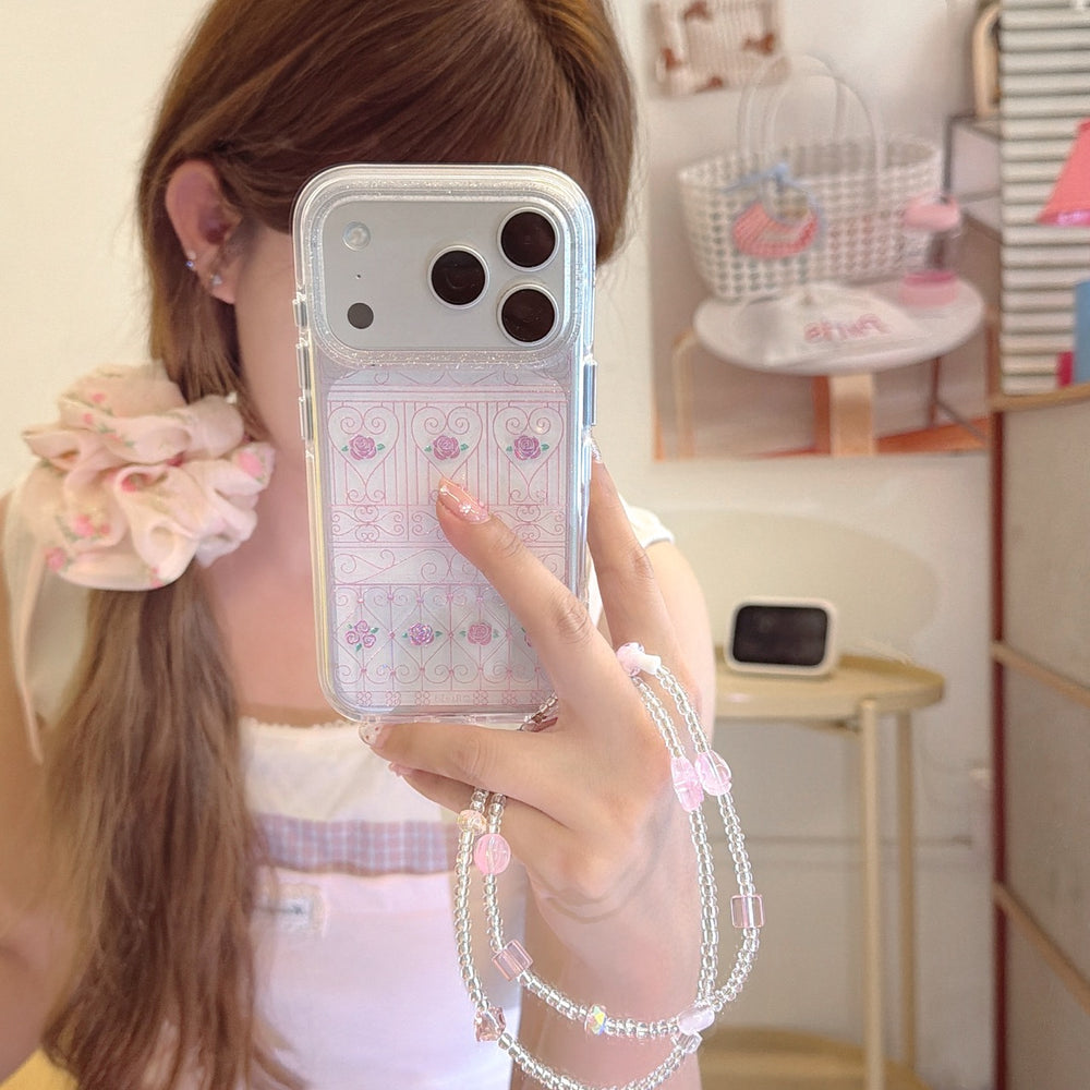 A mirror selfie showing a silver iPhone case with a pink rose gate pattern and a beaded wrist strap.