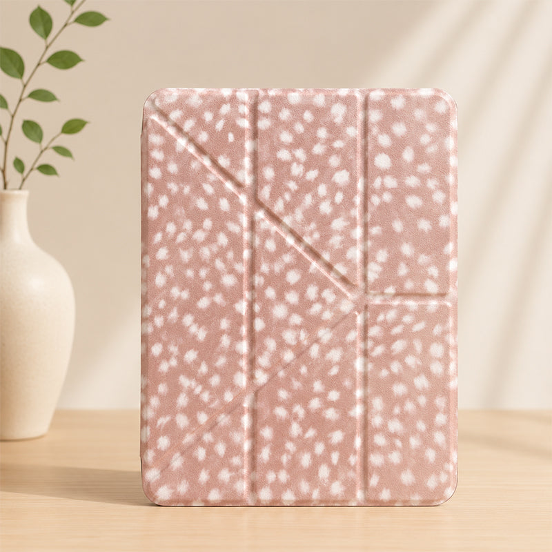 A closed cottagecore deer spot print origami iPad case standing vertically on a wooden table next to a white ceramic vase with green leaves.