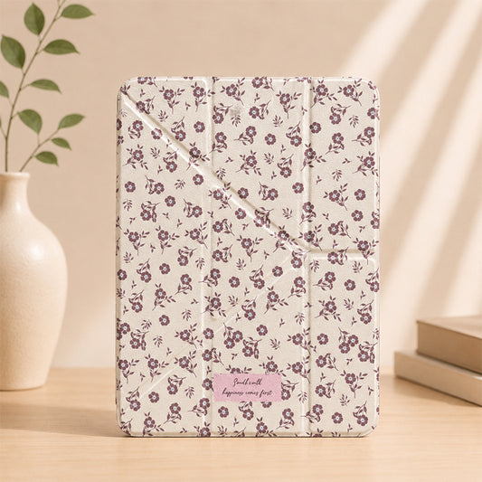 A vertical studio shot of a cream cottagecore floral iPad case standing on a wooden table next to a white ceramic vase with green leaves.