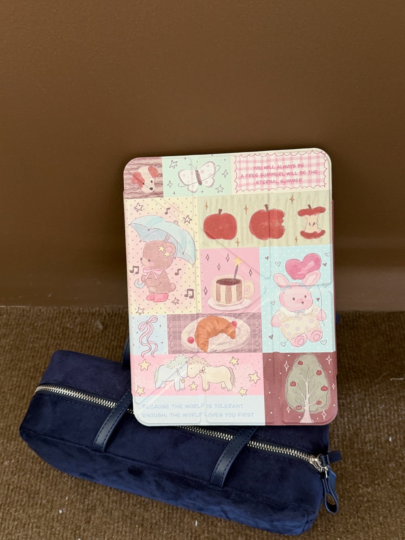 A closed cottagecore teddy bear iPad smart cover leaning vertically against a dark blue velvet pouch on a brown floor.