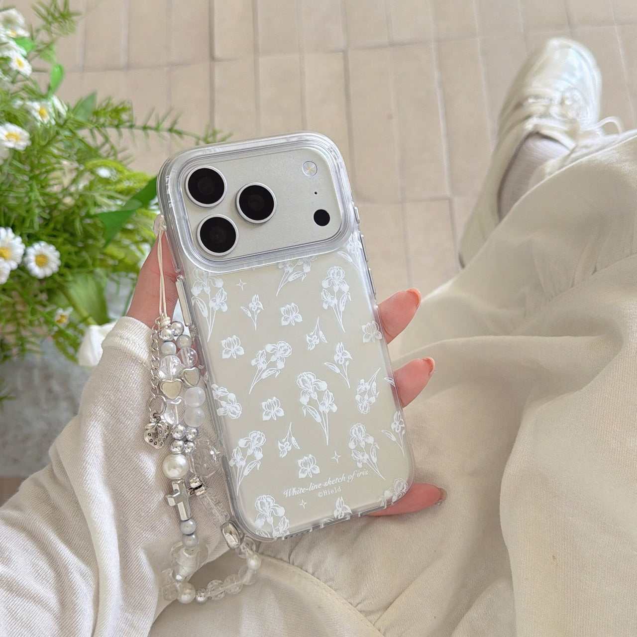 A cottagecore lifestyle photo of a clear white iris phone case held against a cream linen dress near daisies.