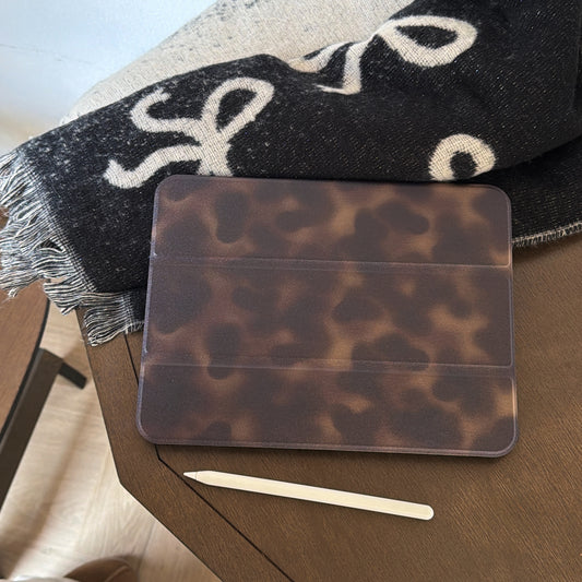 An aesthetic tortoiseshell ipad case lying flat on a wooden desk next to a white Apple Pencil and a black and white patterned bow blanket for a cozy workspace.