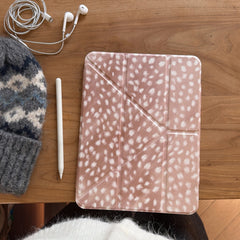 A closed cozy cottagecore deer pattern iPad smart cover lying on a wooden table alongside a white stylus pen, white earphones, and a wool beanie.