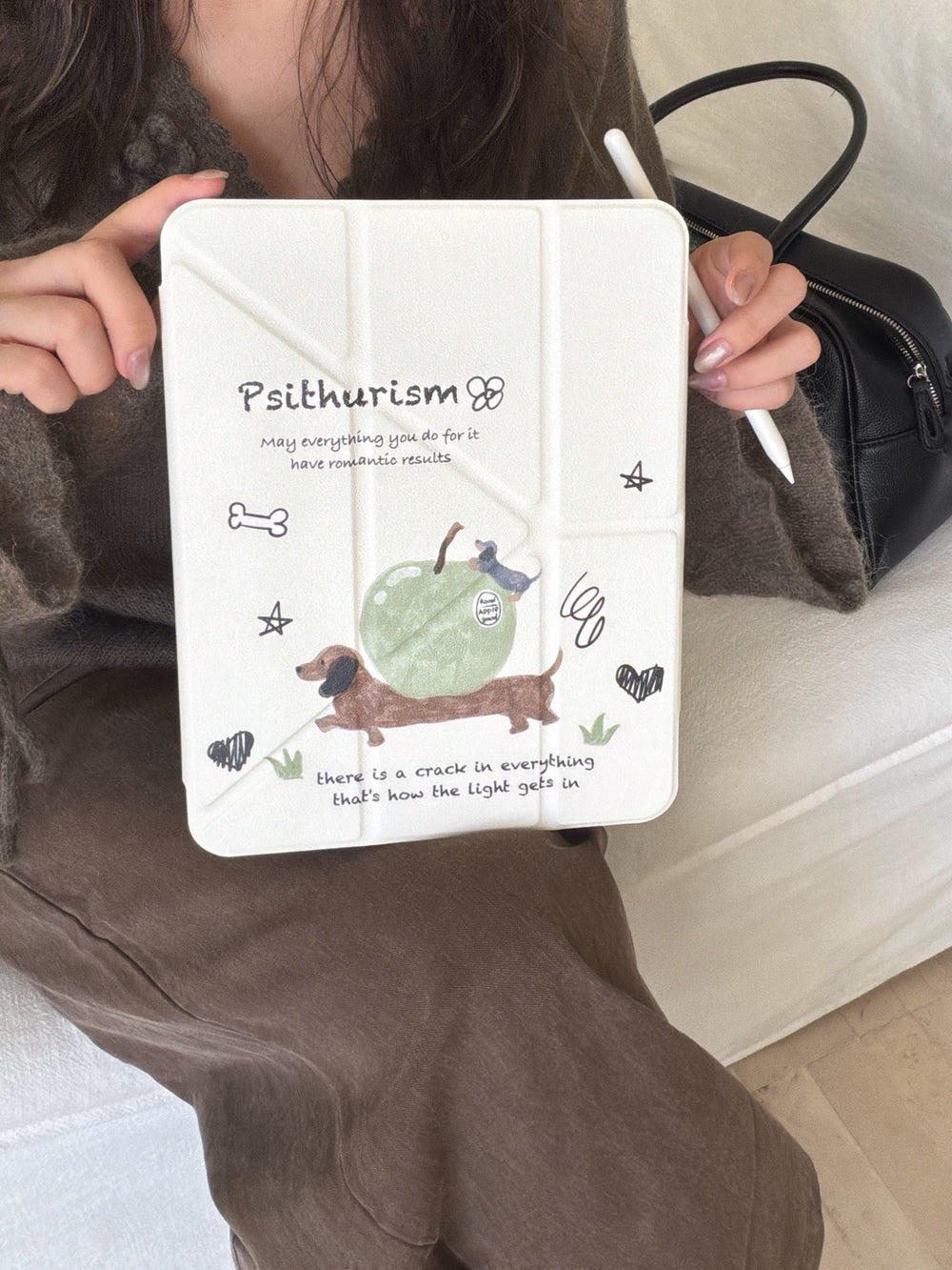 A woman holding a cute aesthetic ipad stand case facing forward, showing the closed cream front cover with the dachshund apple illustration while holding a white Apple Pencil.