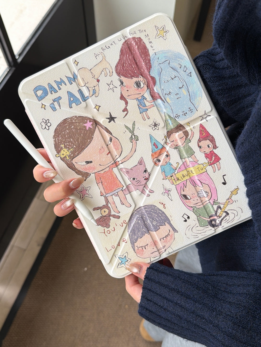 Front view of a cute Yoshitomo Nara art iPad smart case held vertically, showing the cream cover decorated with quirky illustrations and an Apple Pencil.