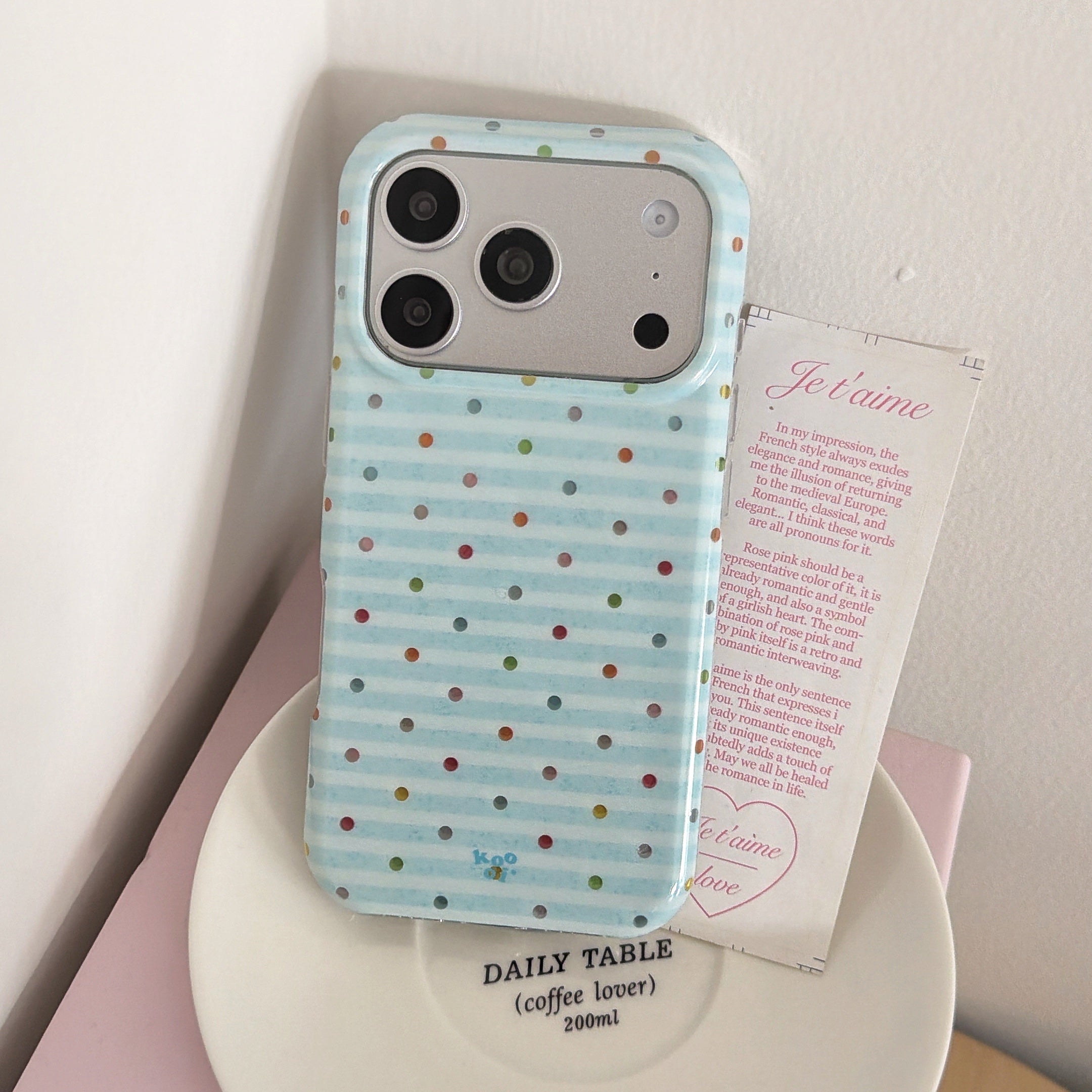 A cute blue polka dot phone case standing on a white ceramic plate next to a romantic French text card.