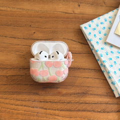 An open cute cherry AirPods case standing upright on a wooden desk next to a blue floral cloth.
