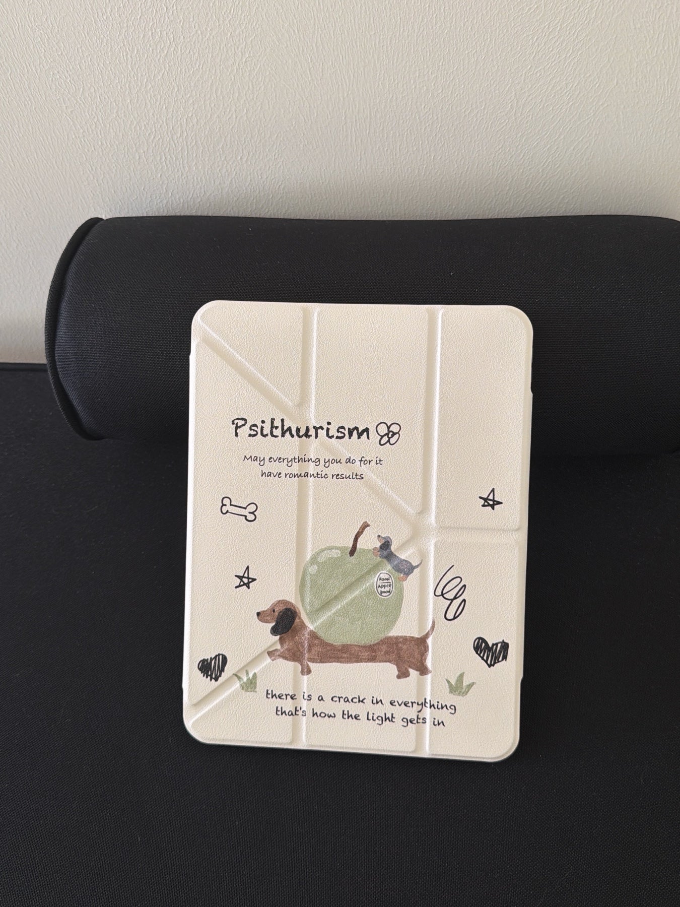 The closed front view of a cute dachshund ipad case standing vertically against a black bolster pillow, highlighting the cream origami cover with hand-drawn dog and apple doodles.