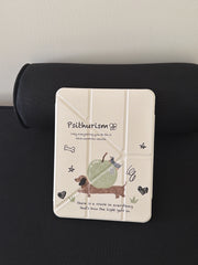 The closed front view of a cute dachshund ipad case standing vertically against a black bolster pillow, highlighting the cream origami cover with hand-drawn dog and apple doodles.