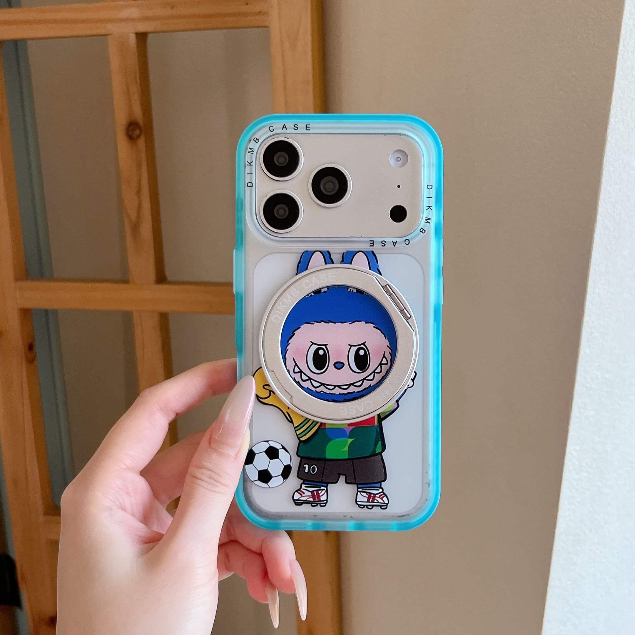 A hand with manicured nails holding a cute Labubu soccer phone case with a clear mint blue bumper.