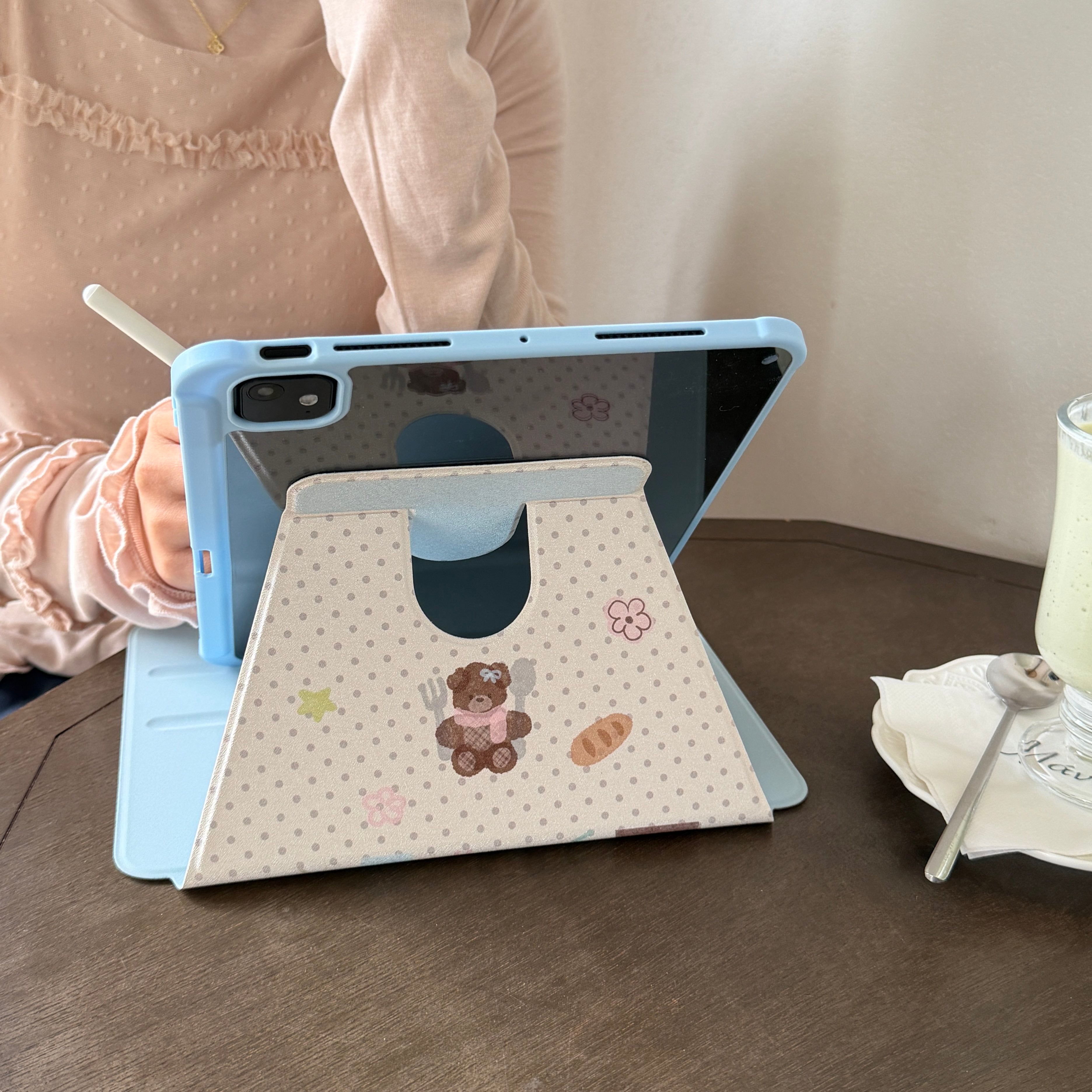 An aesthetic cafe desk setup featuring a cute pastel blue iPad case standing stably on a wooden table using its folded origami cover, next to a refreshing iced matcha latte.