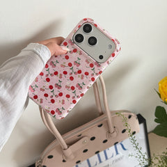 A hand holding a cute pink cherry phone case over a beige polka dot handbag and a yellow rose.