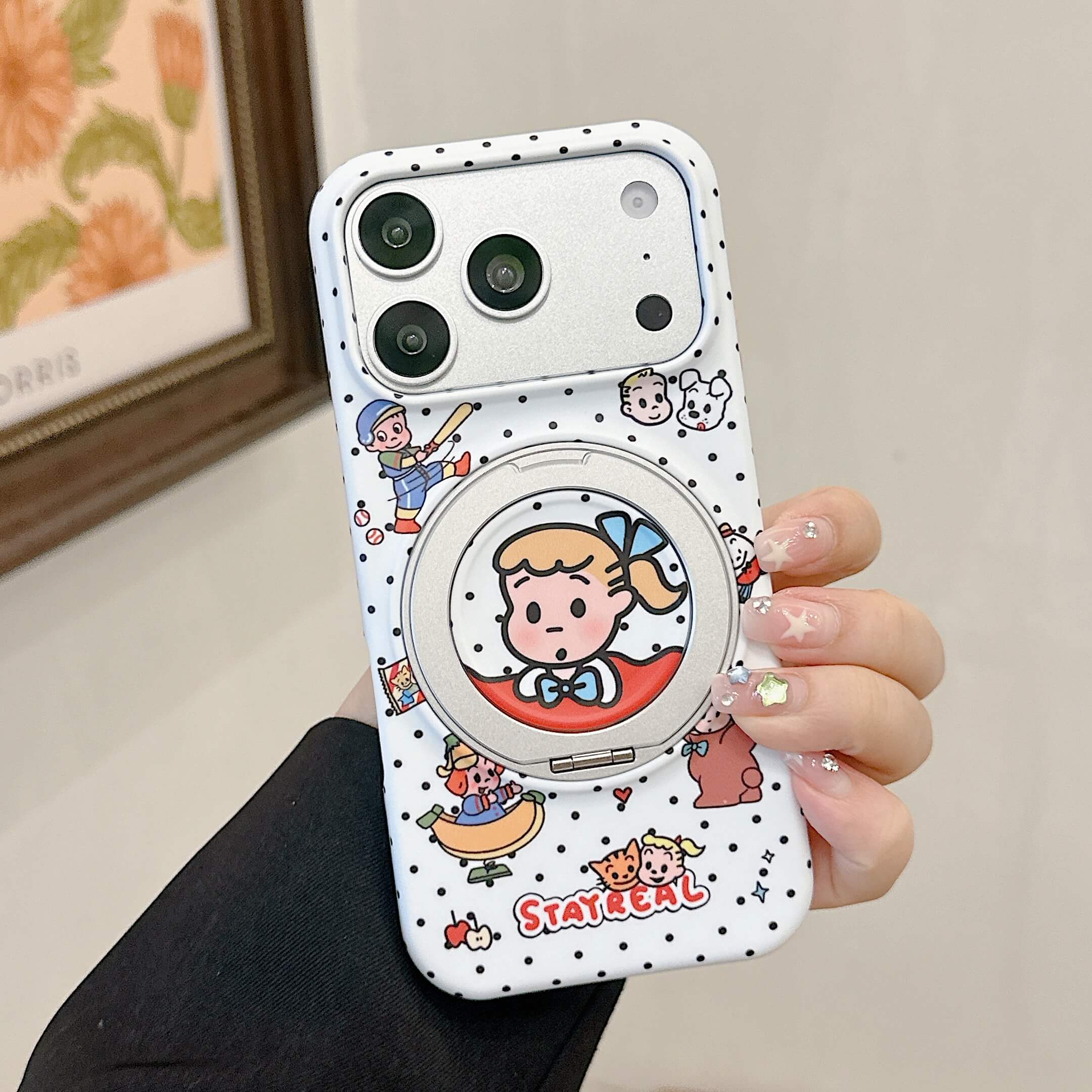 A woman's hand holding a white polka dot iPhone case featuring retro cartoon characters and a folded silver magnetic stand.