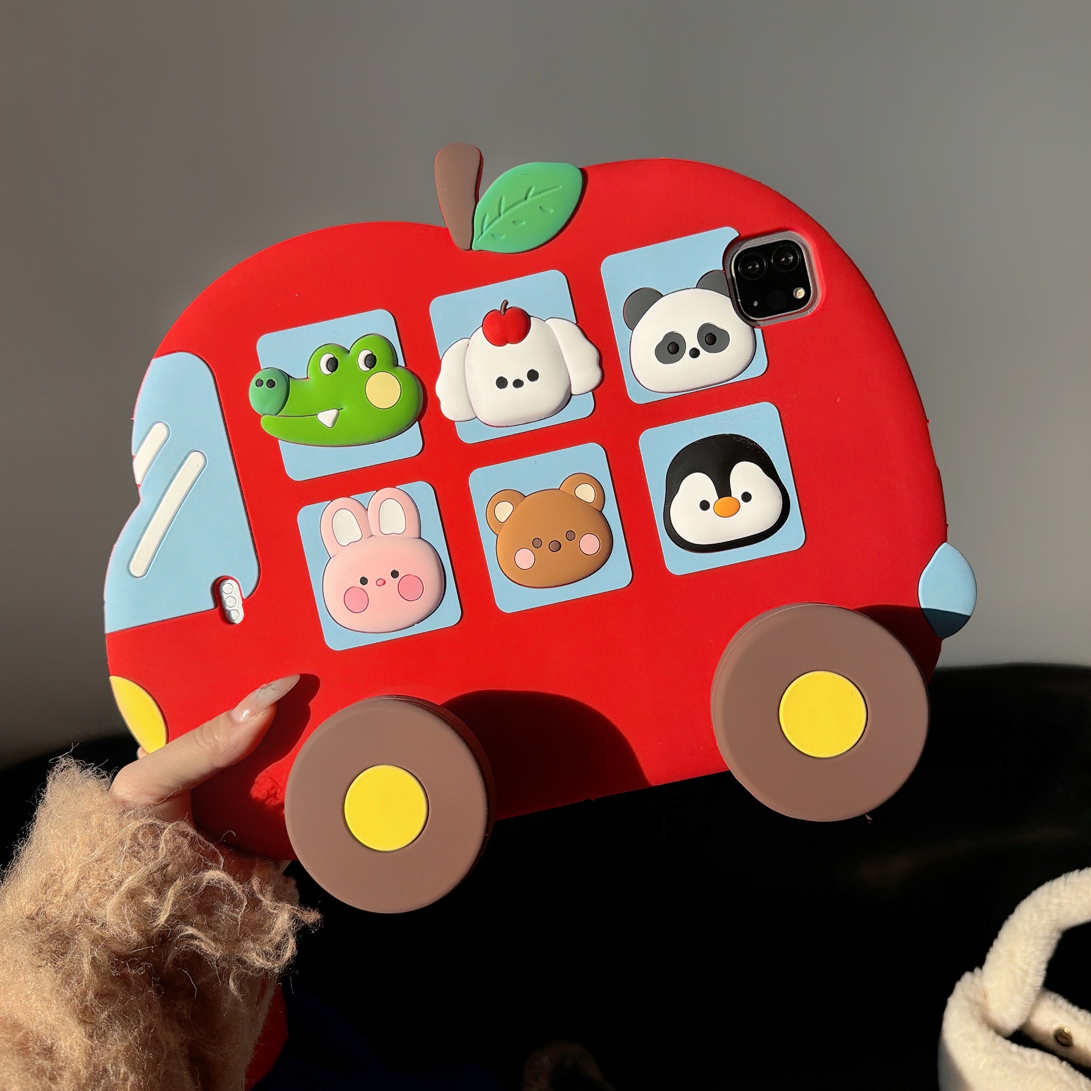 A hand holding the cute red bus iPad case vertically under bright sunlight, highlighting the vivid colors of the 3D animal passengers, the apple shape, and the camera cutout.
