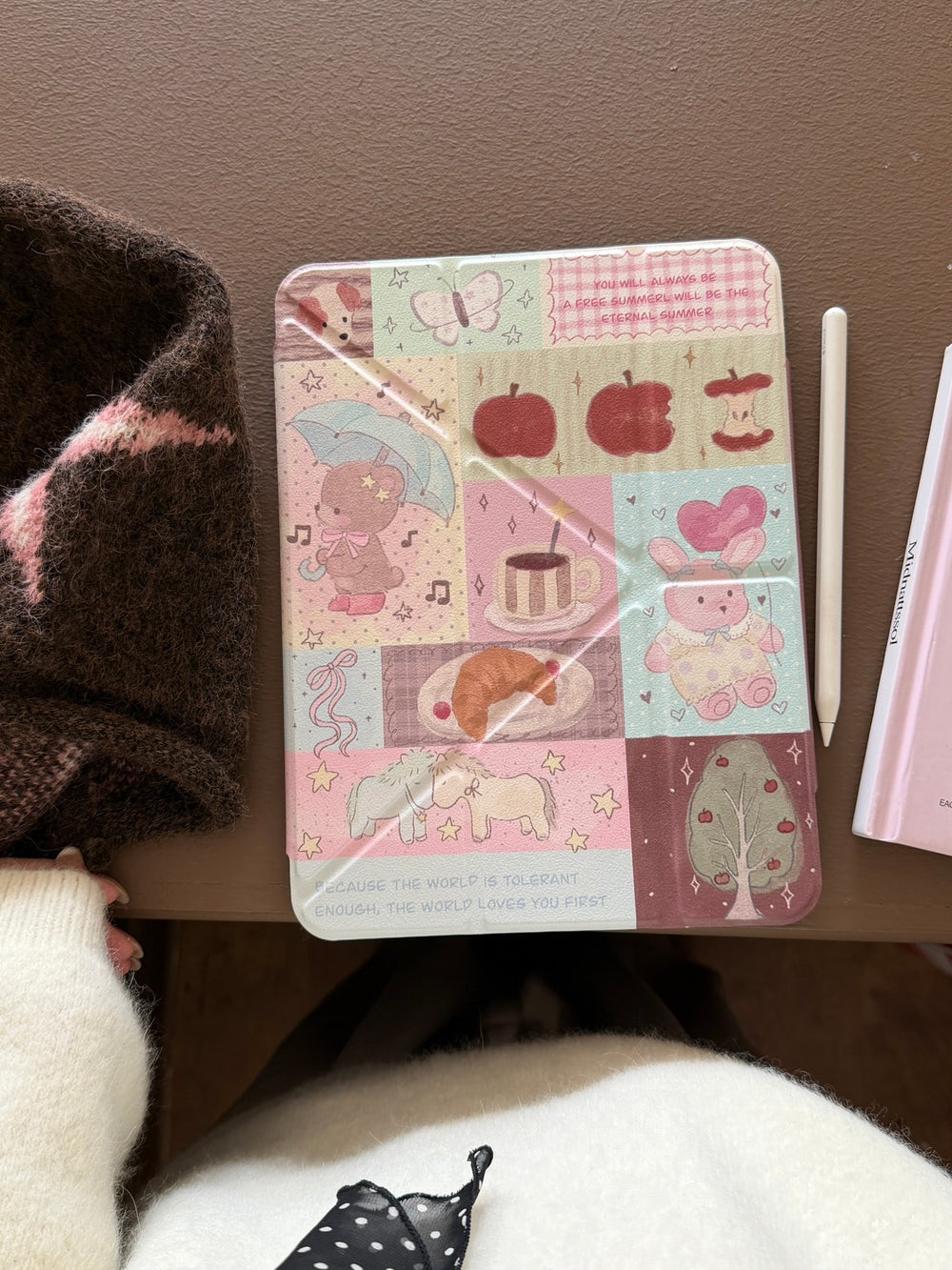 Top-down flat lay view of a closed cute teddy bear patchwork iPad case on a brown table next to a white stylus pencil.