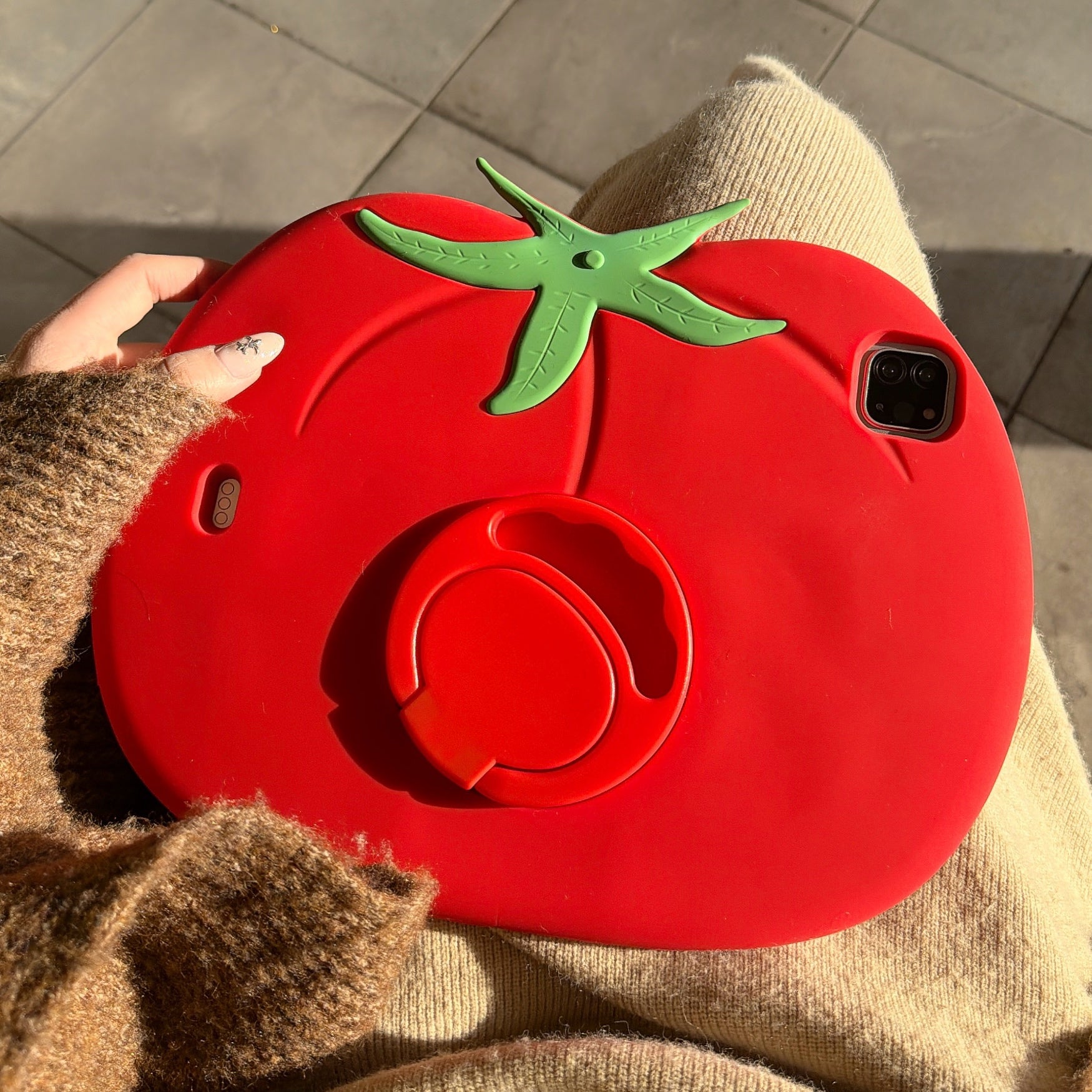 A person holding a cute tomato ipad case on their lap in direct sunlight, showcasing the full back design with its green leaf topper and folded circular ring kickstand.