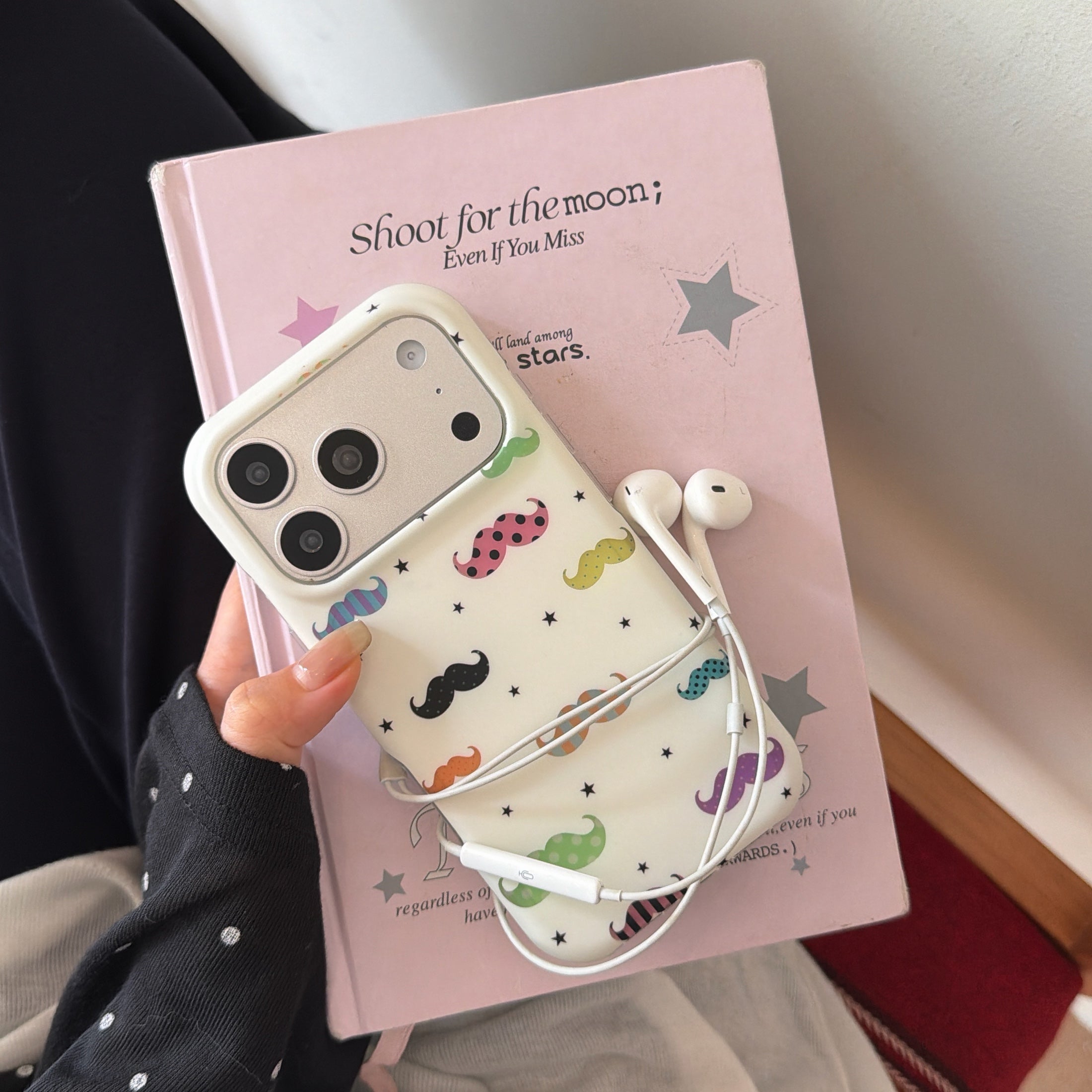A hand holding a white mustache phone case on a pink book with wrapped white earphones.