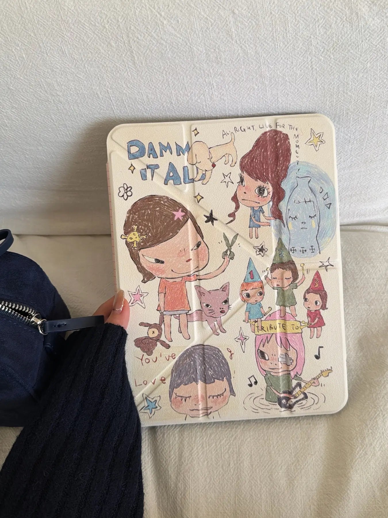 Close-up of the front cover of a Yoshitomo Nara iPad case featuring cute whimsical hand-drawn art, cartoon girls, and "DAMN IT ALL" text doodle.