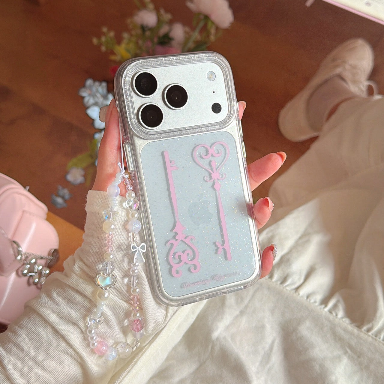 A person in a white outfit holding a fairycore glitter phone case with a pink key graphic and beaded strap.