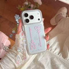 A person in a white outfit holding a fairycore glitter phone case with a pink key graphic and beaded strap.