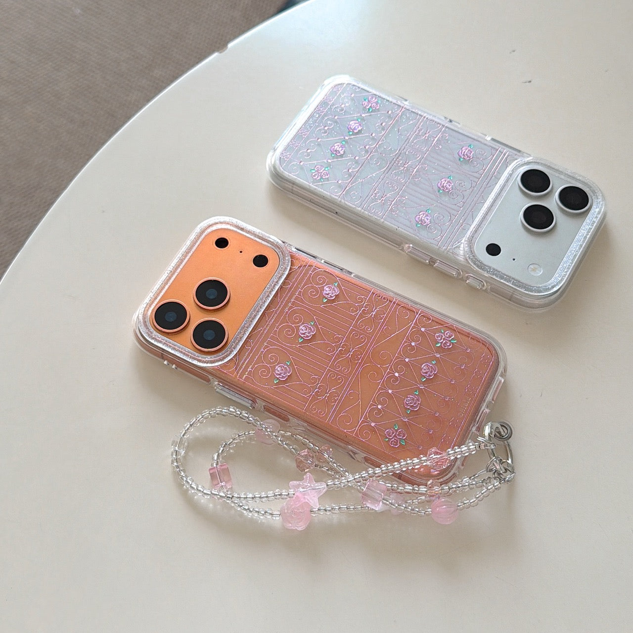 Two clear glitter phone cases with pink rose fence designs on a white table.