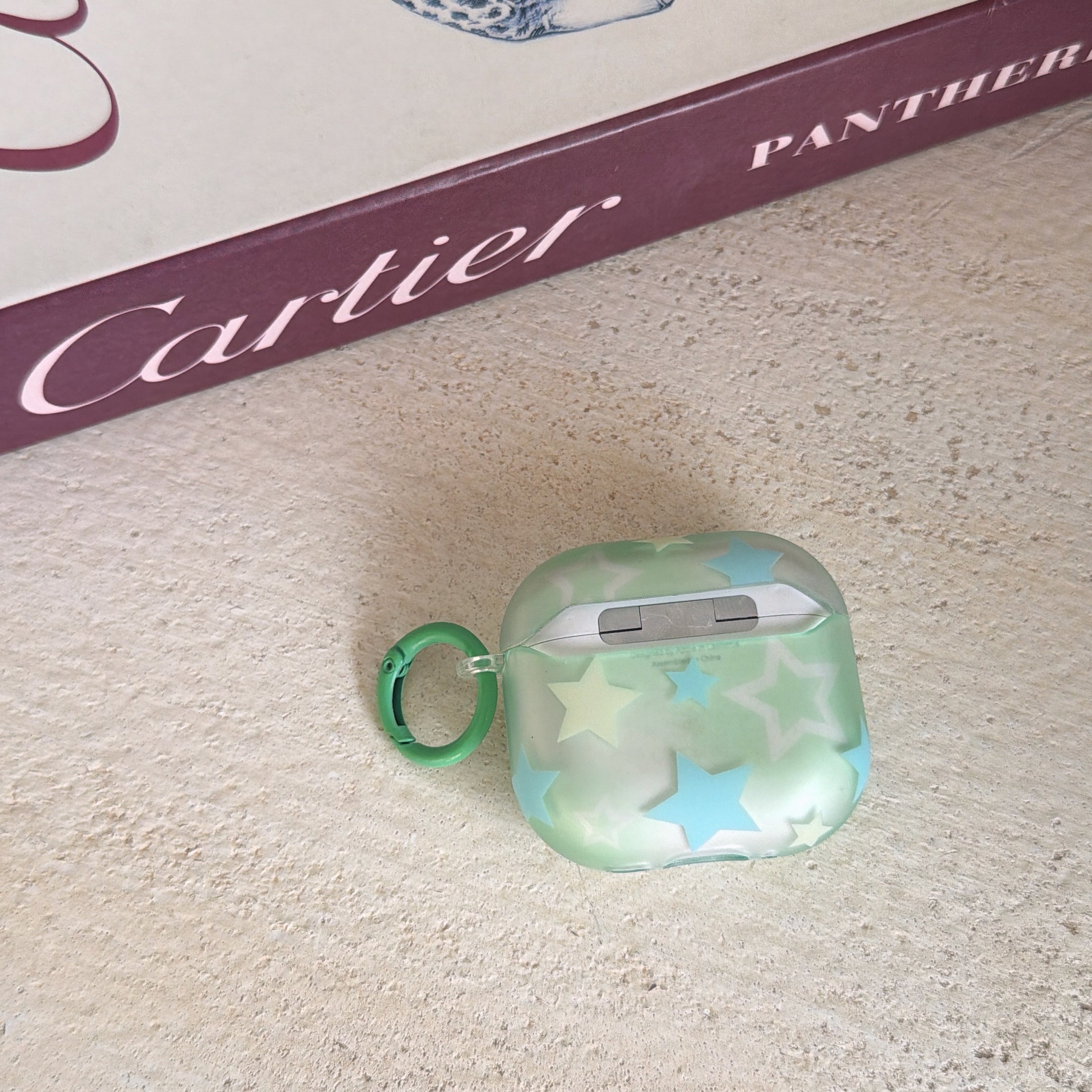 The back view of a frosted mint green starry AirPods case with a green ring carabiner resting below a Cartier book.
