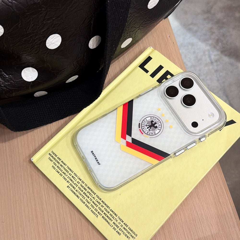 A silver Germany 4-stars mirror phone case lying flat on a yellow book next to a polka dot bag.