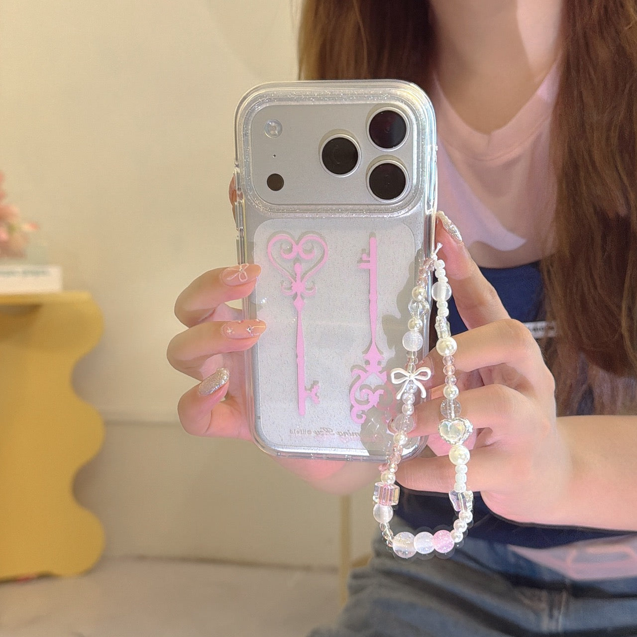 Close-up of a hand holding a clear glittery magic key phone cover featuring a pastel beaded lanyard with a white bow.