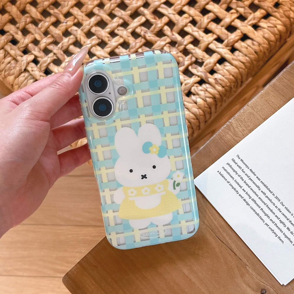 A hand holding a pastel blue plaid cartoon bunny iPhone case over a wooden surface.
