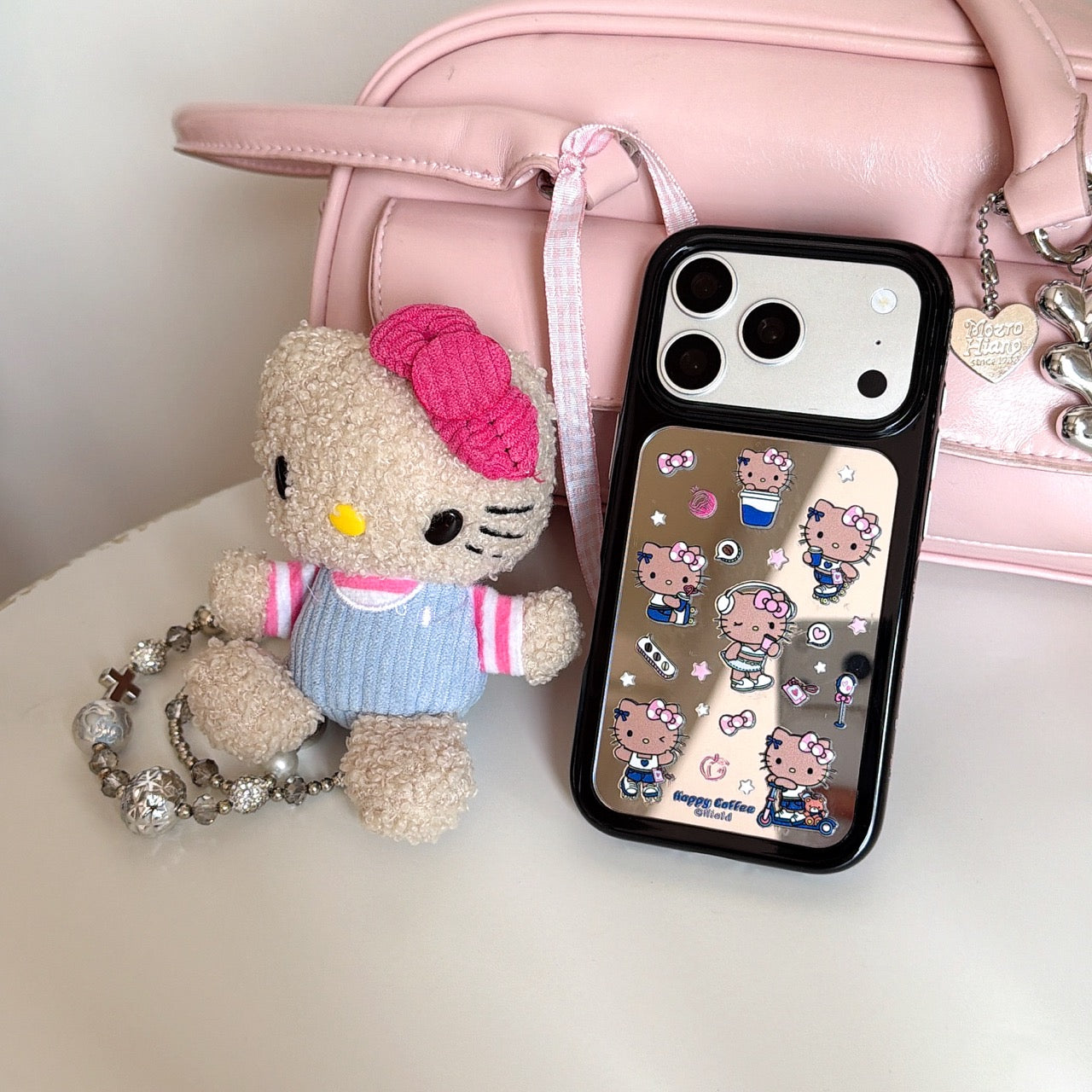 A silver Hello Kitty mirror iPhone case displayed on a white table next to a matching plush doll and a pastel pink handbag.