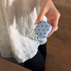A close-up of a hand holding a light blue polka dot AirPods case against a white embroidered dress.
