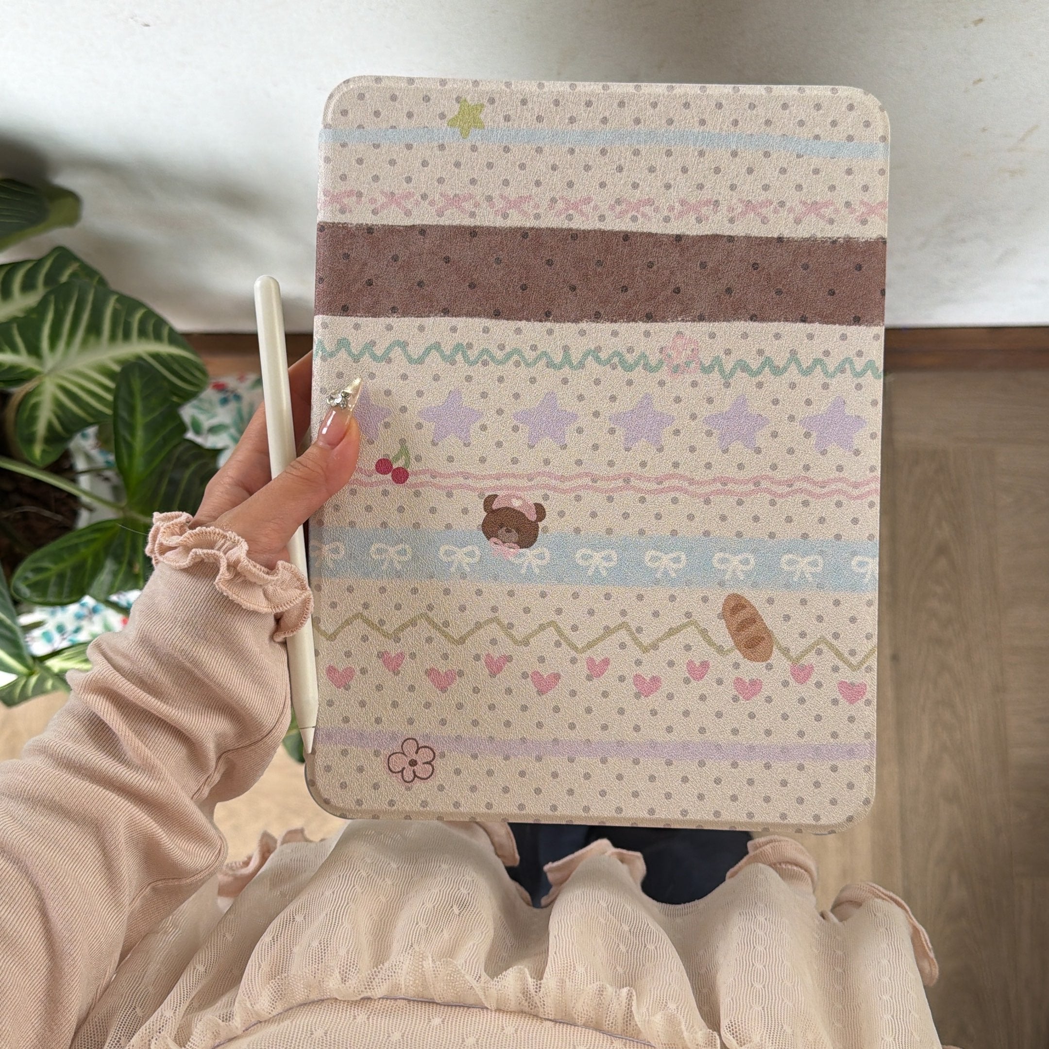 Top-down shot of a girl holding a closed kawaii teddy bear iPad case vertically, showcasing the complete colorful scrapbook pattern with stars, bows, and an attached white Apple Pencil.